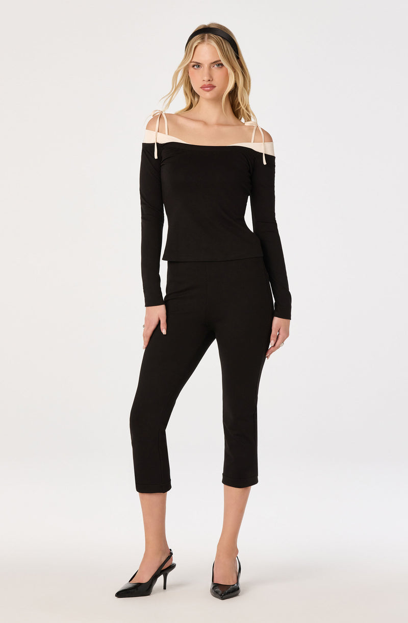 Woman in black off-shoulder top with white shoulder straps and matching black pants, wearing black heels.