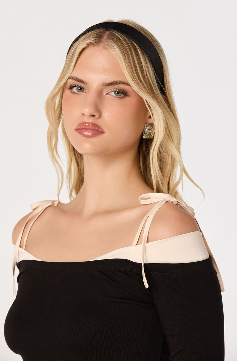 Woman with blonde hair wearing a black headband and an off-the-shoulder black and white top with beige shoulder ties.