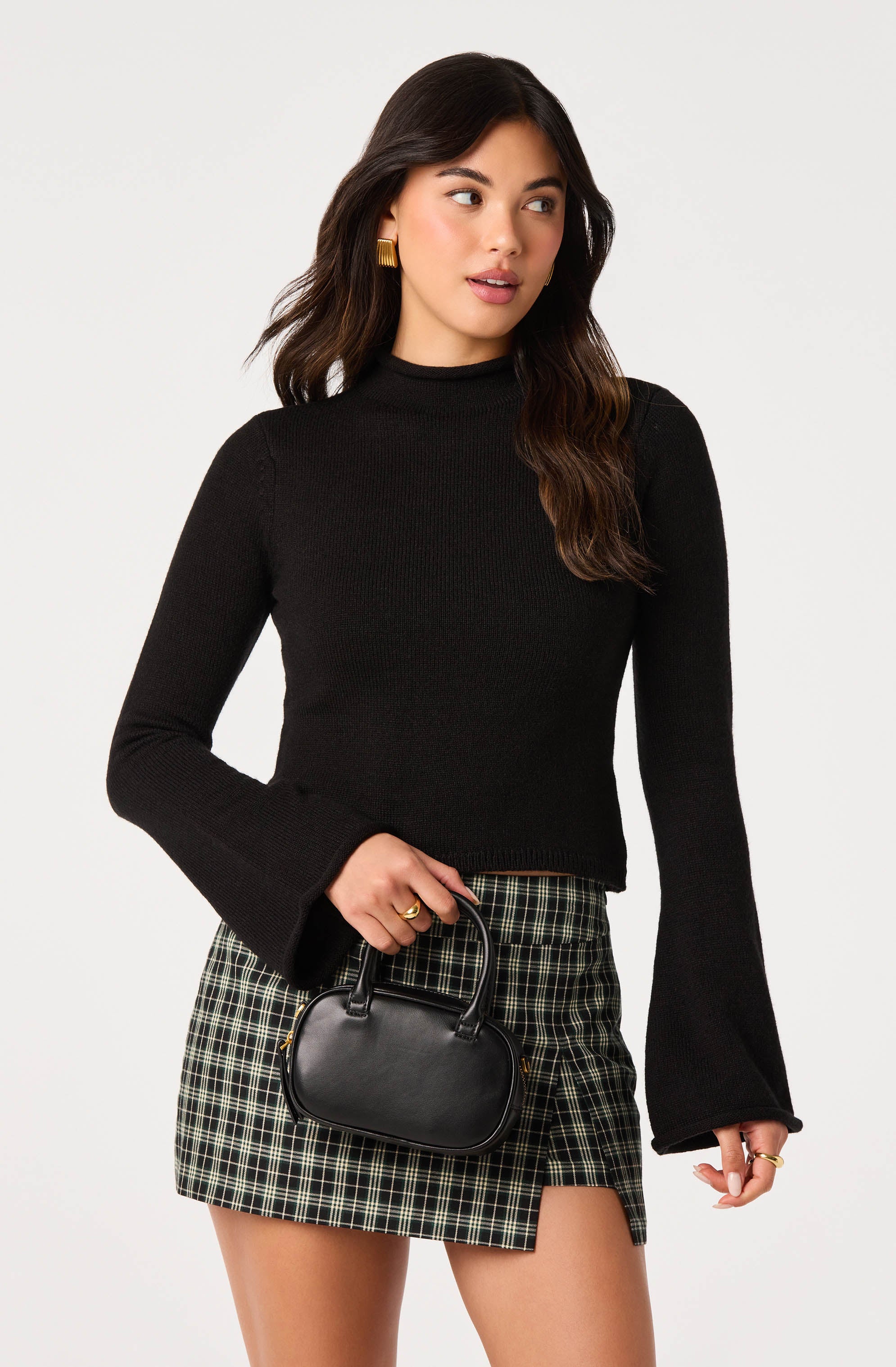 Woman wearing a black long-sleeve top with bell sleeves and plaid skirt, holding a small black handbag.