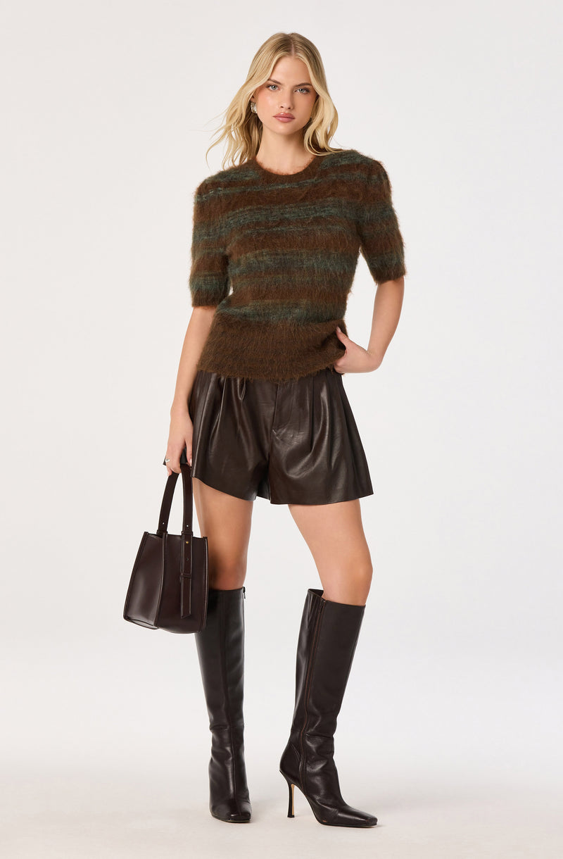 Woman wearing a multicolor fuzzy short-sleeve sweater, black leather shorts, and tall black boots, holding a black handbag.