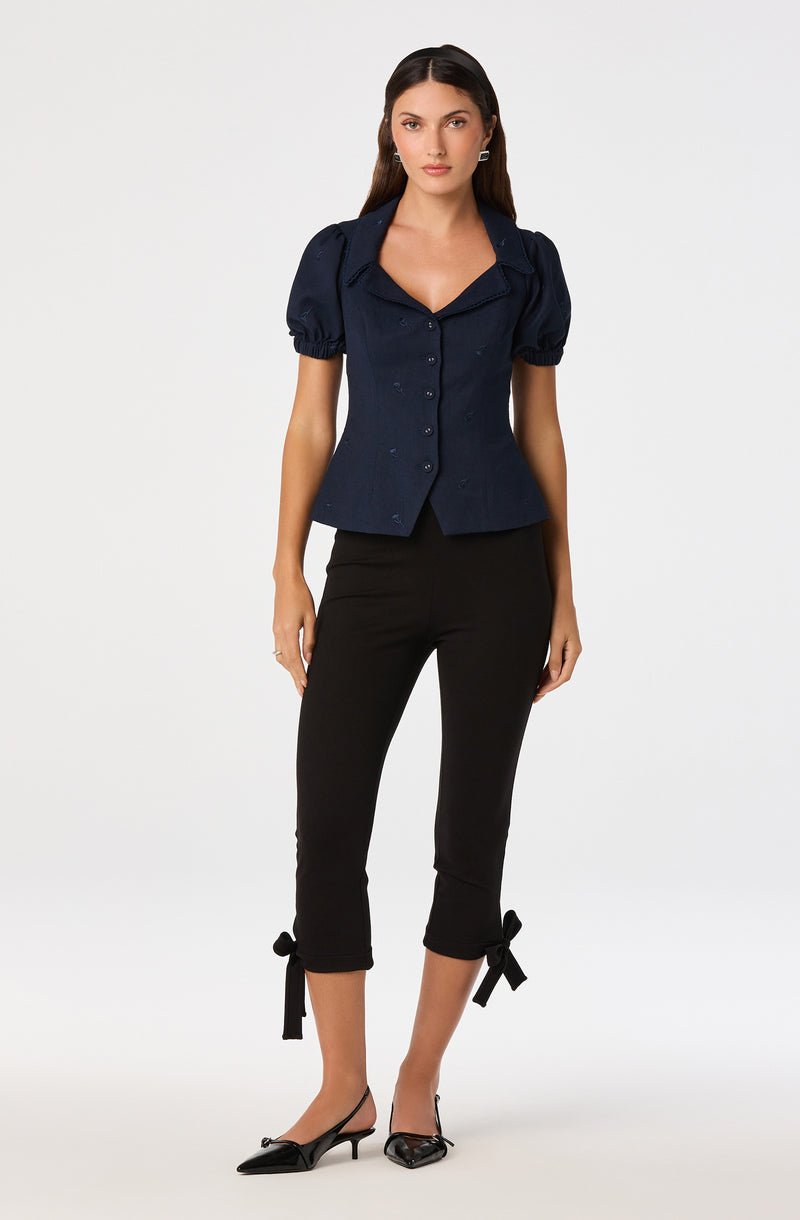 Woman wearing a navy blue short-sleeve blouse with puffed sleeves and black cropped pants with bow ties.