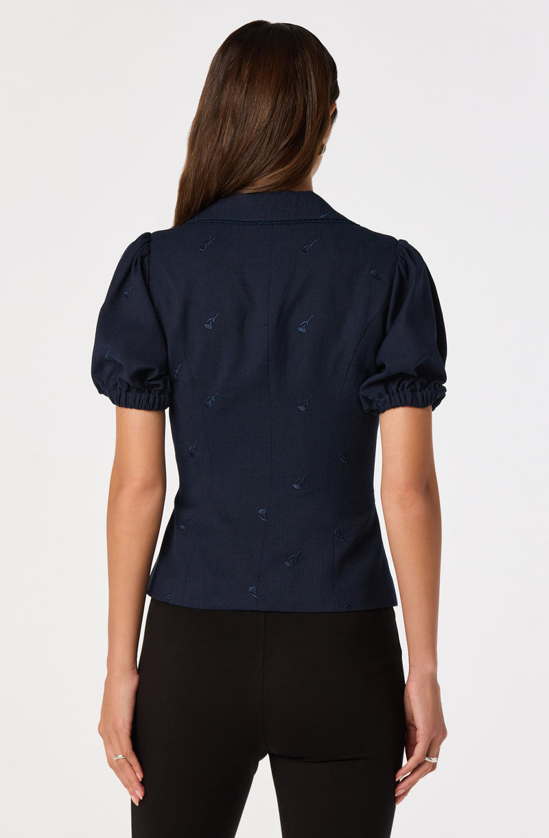 Woman wearing a navy blue blouse with puffed sleeves and embroidered details, paired with black pants.