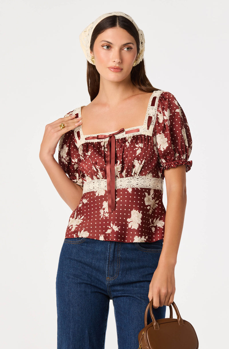 Woman wearing a maroon floral and polka dot blouse with lace details and puffed sleeves, paired with blue jeans and a cream-colored headscarf.