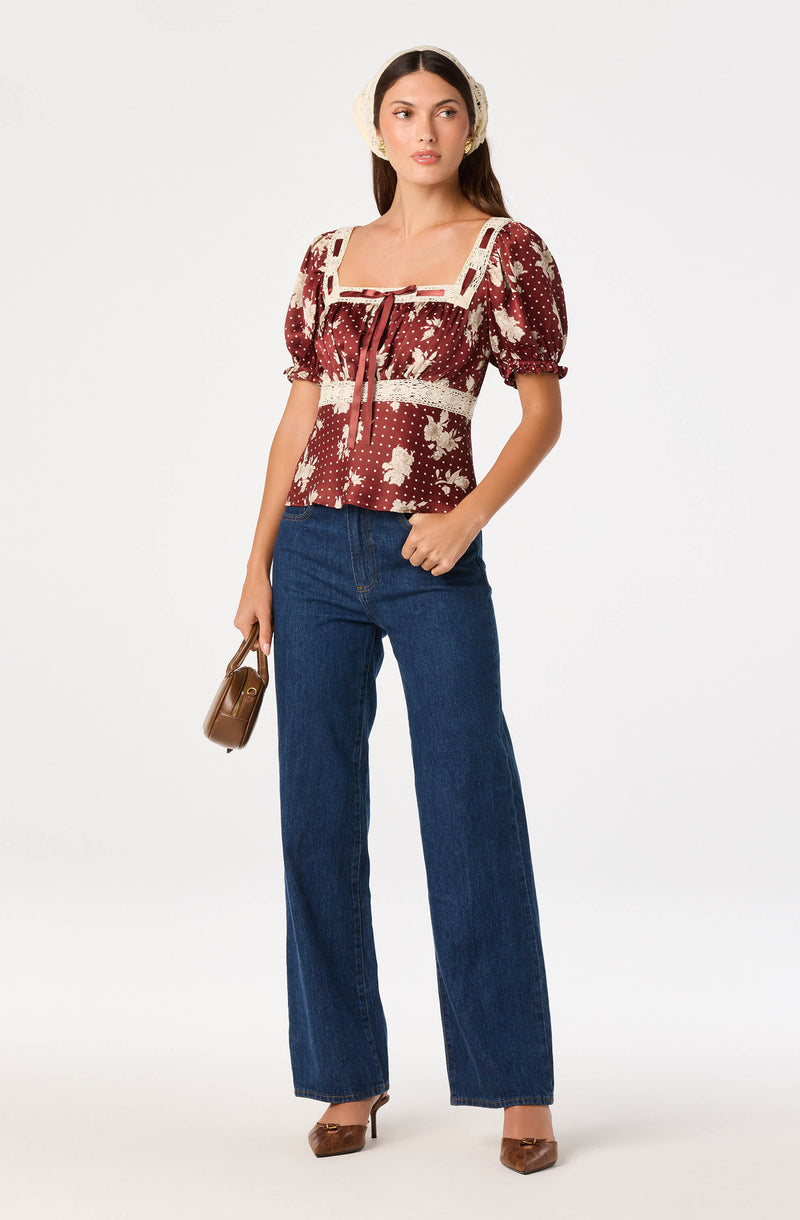 Woman wearing a maroon floral blouse with puffed sleeves, beige lace details, high-waisted wide-leg jeans, beige high heels, and beige earmuffs.