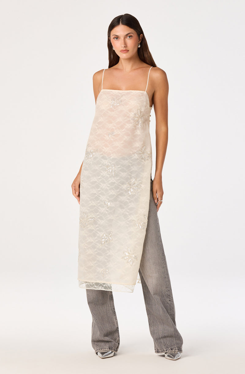 Woman wearing a sheer, cream-colored lace slip dress with spaghetti straps over gray wide-leg jeans.
