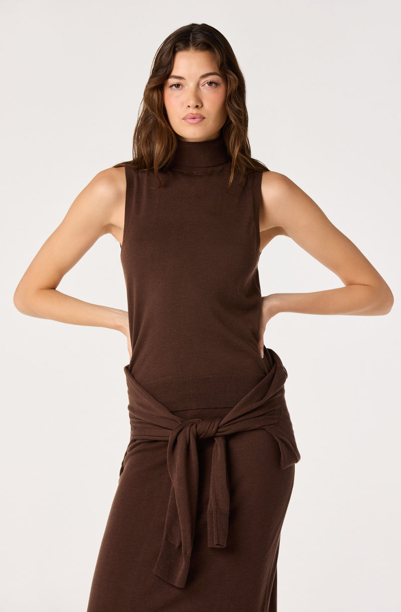 Woman wearing a sleeveless brown turtleneck top and a matching brown skirt with a tied sweater around her waist.