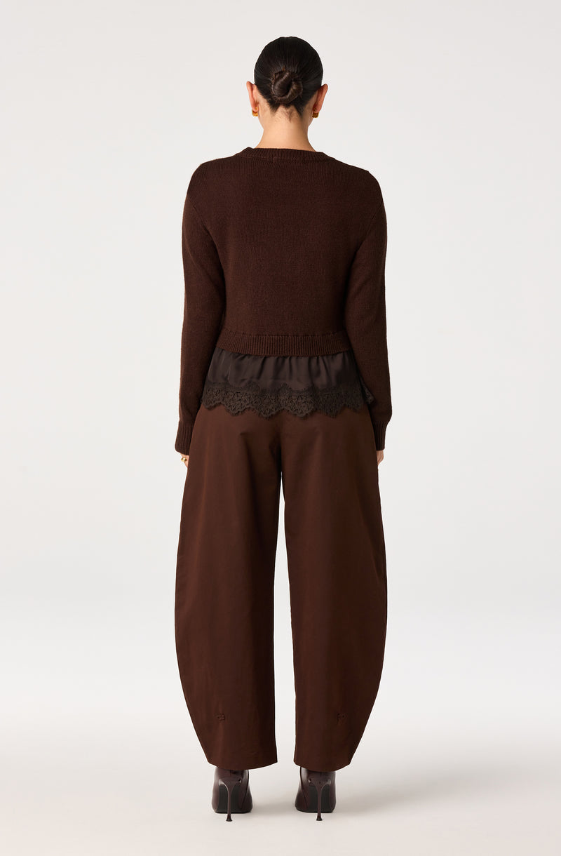Woman wearing a brown sweater, dark brown lace-trimmed top, wide brown pants, and high-heeled shoes.