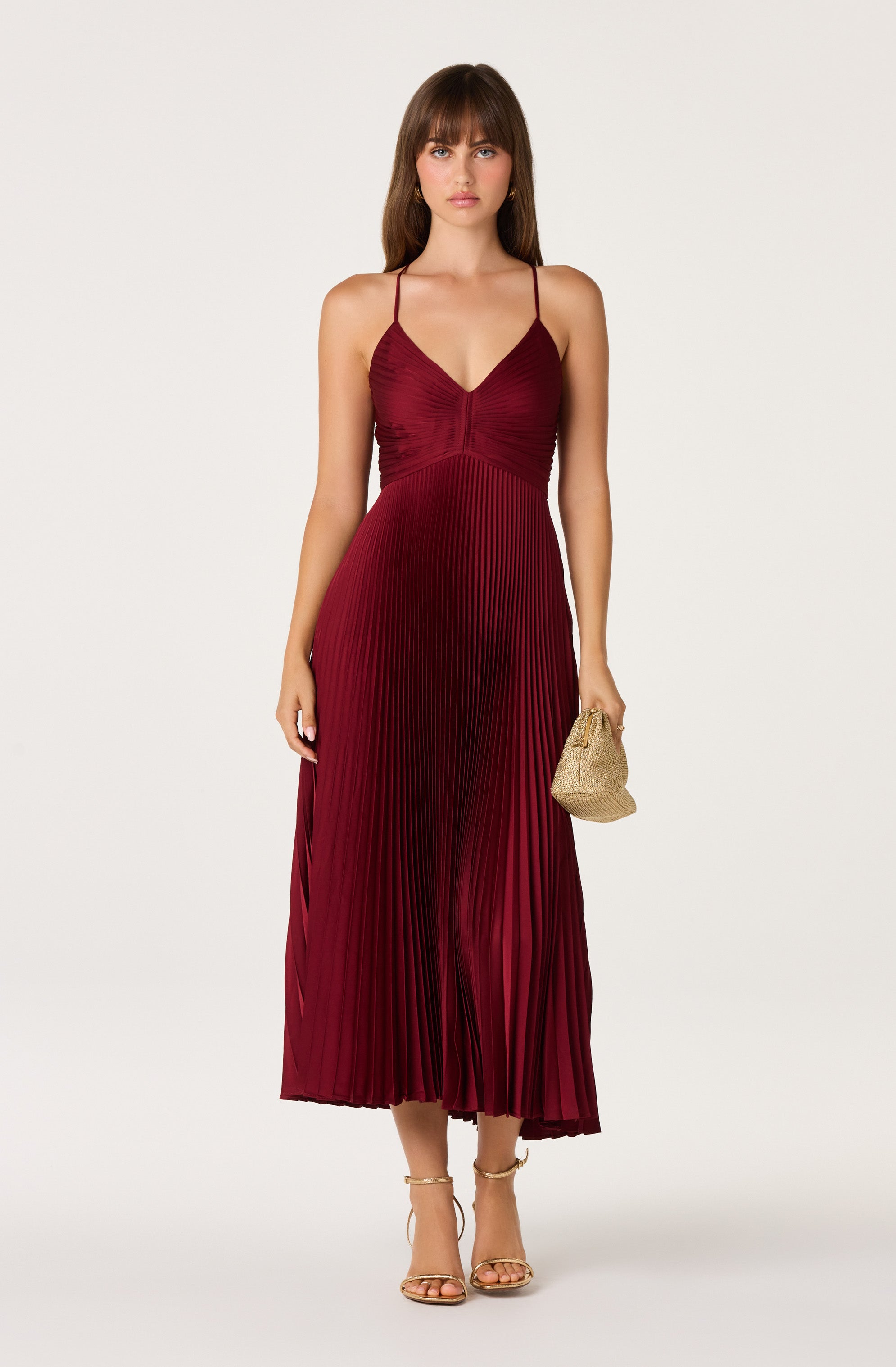 Woman in a burgundy pleated midi dress with spaghetti straps, holding a beige clutch.