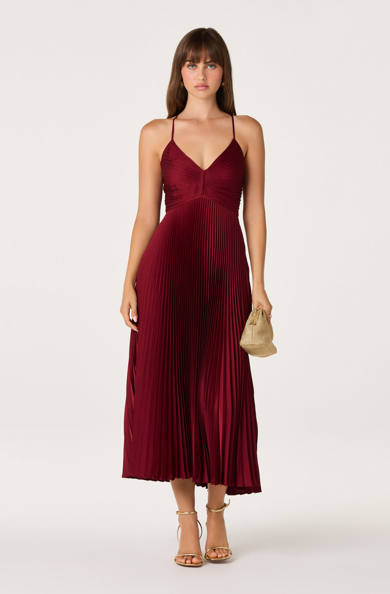 Woman in a burgundy pleated midi dress with spaghetti straps, holding a beige clutch.