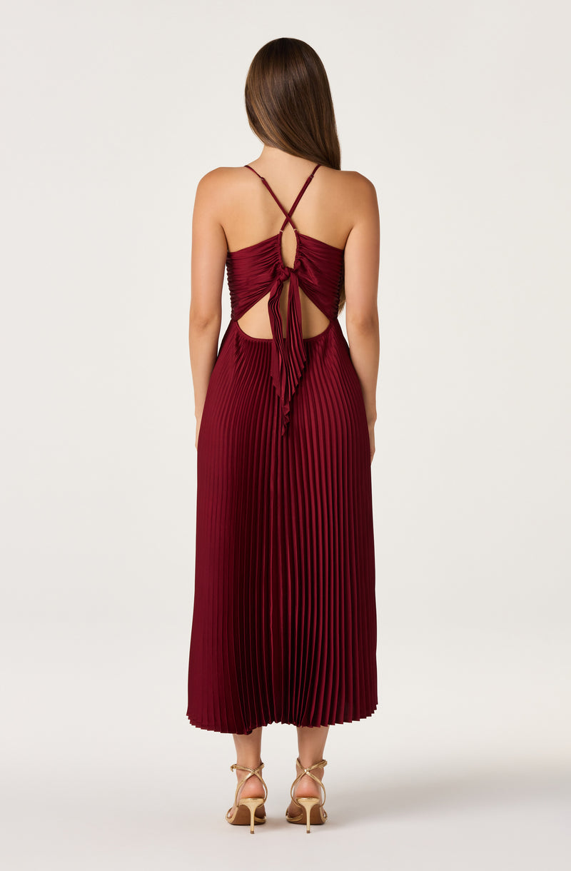 Back view of a woman wearing a deep red, pleated dress with a ruched, knotted back and thin straps.