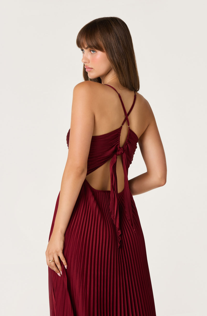 Woman wearing a sleeveless, pleated, deep red dress with thin, crossed straps and a back tie.