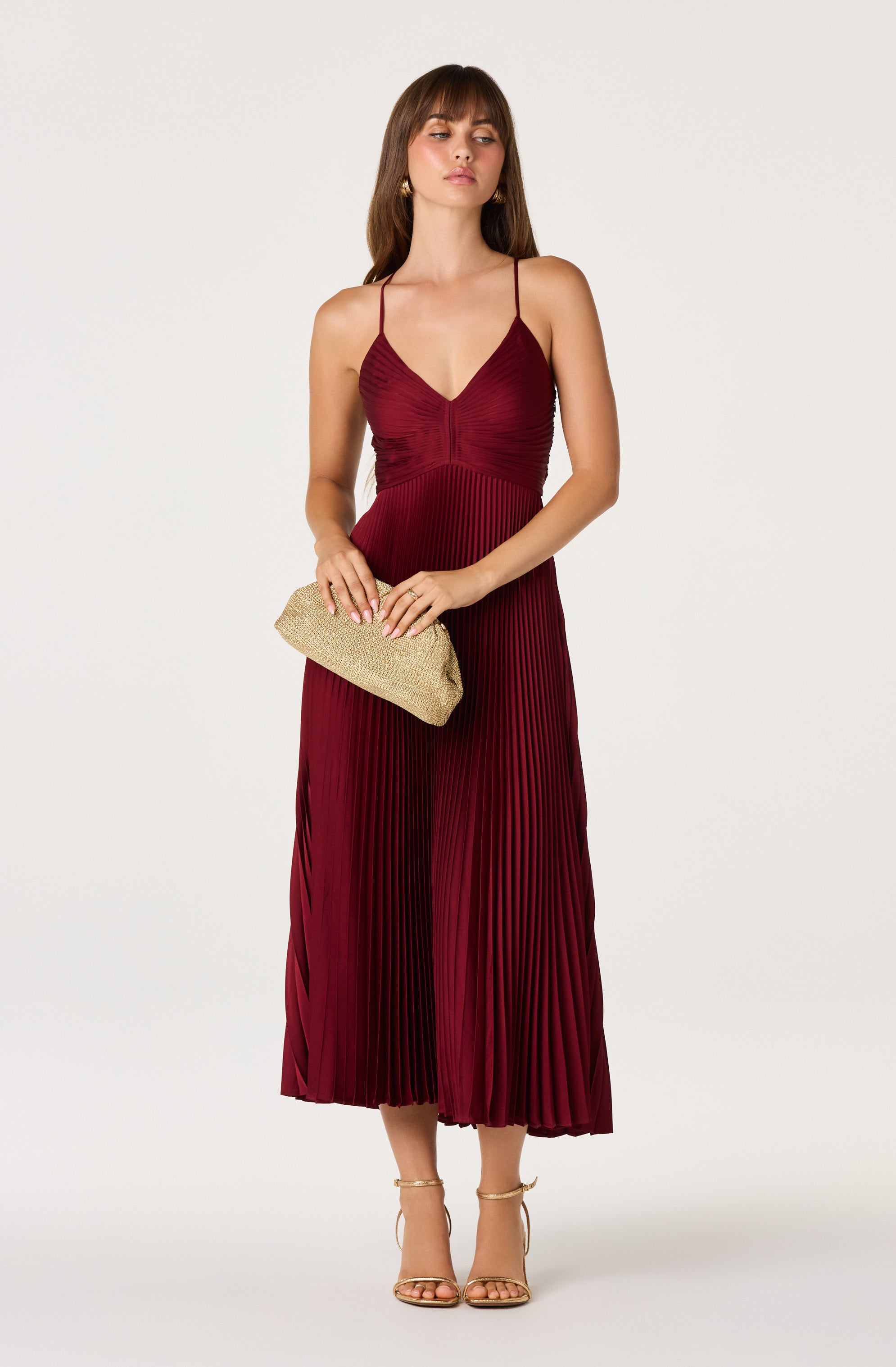 Woman in a long, pleated, burgundy dress with spaghetti straps, holding a beige clutch.