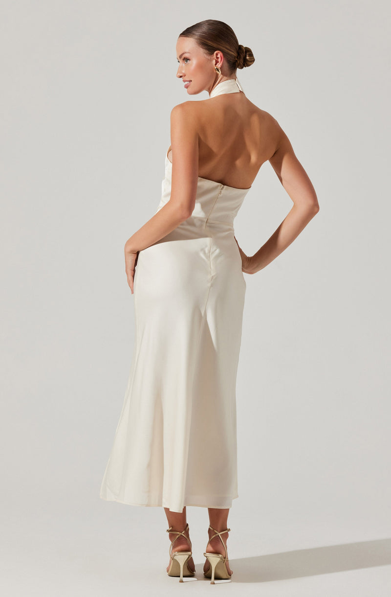 A woman is wearing a long, elegant satin dress in a creamy white color with a halter neckline that reveals her back. The dress has a fitted waist and flows down to her ankles. She is accessorized with gold hoop earrings and beige strappy high heels.