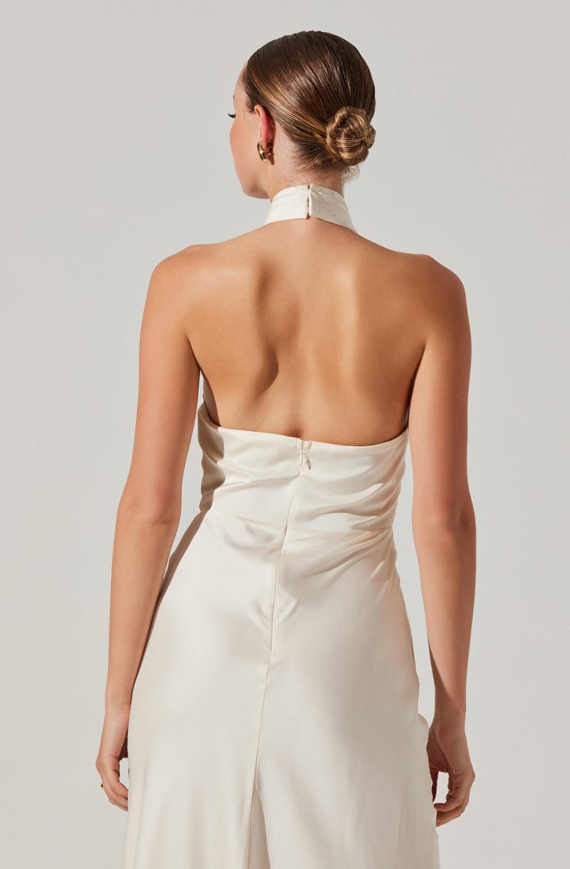 A woman wearing a sleeveless, backless, ivory satin dress with a high neck and a zipper at the back. Her hair is styled in a sleek, low bun, and she has gold hoop earrings.