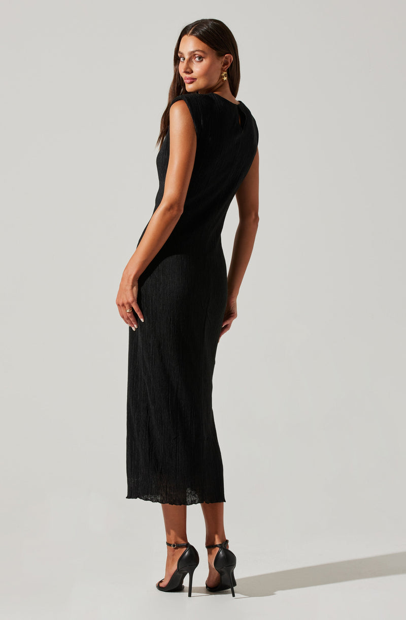The woman is wearing a sleeveless, black, ankle-length dress made of a shiny, ribbed fabric. She has on black high-heeled shoes with ankle straps. Her hairstyle is long, dark, and straight. She accessorizes with gold earrings and a ring on her left hand.