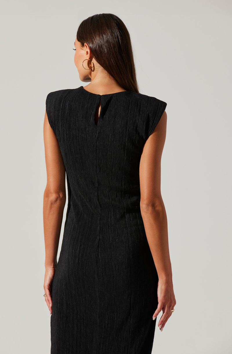 A woman is wearing a sleeveless, black ribbed dress with a small keyhole opening at the back near the neckline. The dress has structured shoulders and a fitted silhouette. She is accessorized with gold hoop earrings and a ring on her right hand.