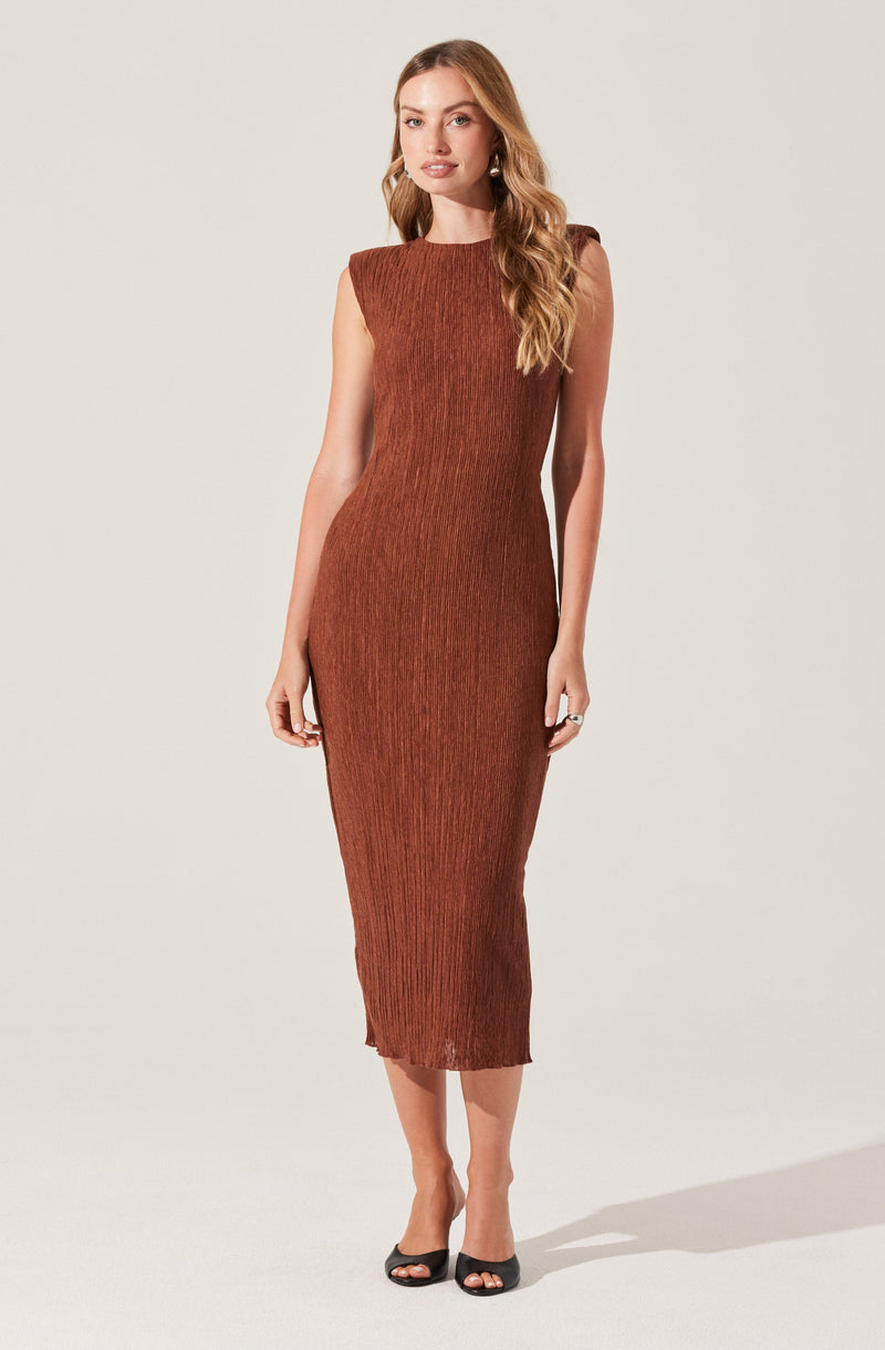 A woman is wearing a sleeveless, form-fitting, knee-length dress in a rust color with a textured, pleated pattern. She has long, wavy blonde hair and is wearing black open-toe heels.