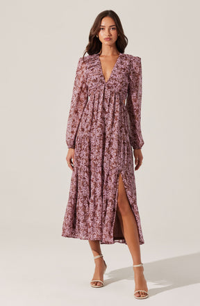 A woman is wearing a long, mauve-colored, patterned dress with long sleeves and puffed shoulders. The dress has a deep V-neckline and features a high slit on one side, revealing her leg. She is also wearing strappy high-heeled sandals.