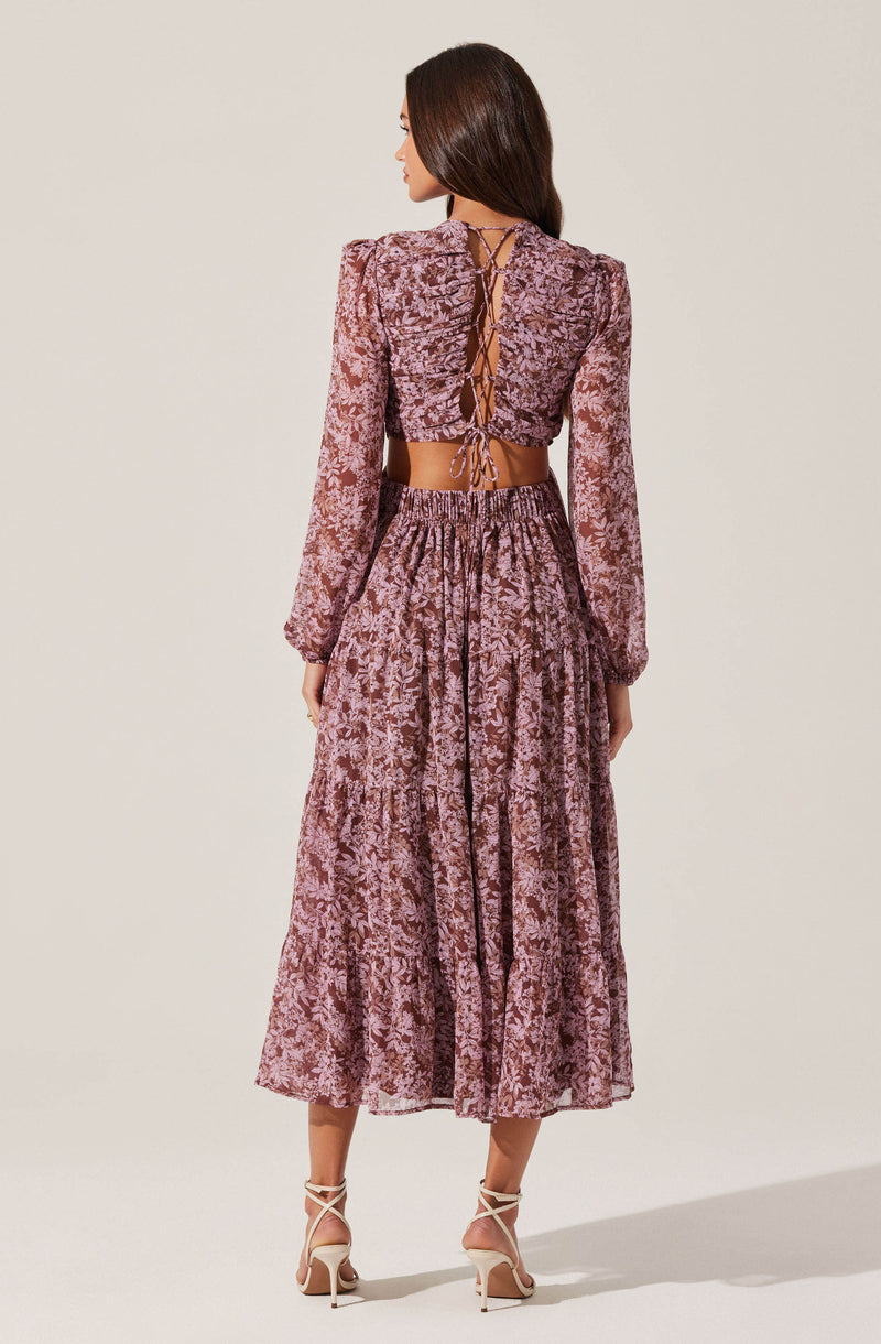 A woman is wearing a two-piece outfit made of pink floral-printed fabric. The top is a long-sleeved, cropped blouse with puffed shoulders and a lace-up back detail tied with a bow. The skirt is a high-waisted, ankle-length maxi with an elastic waistband and a tiered, ruffled design. She is also wearing nude-colored, strappy high-heeled sandals.