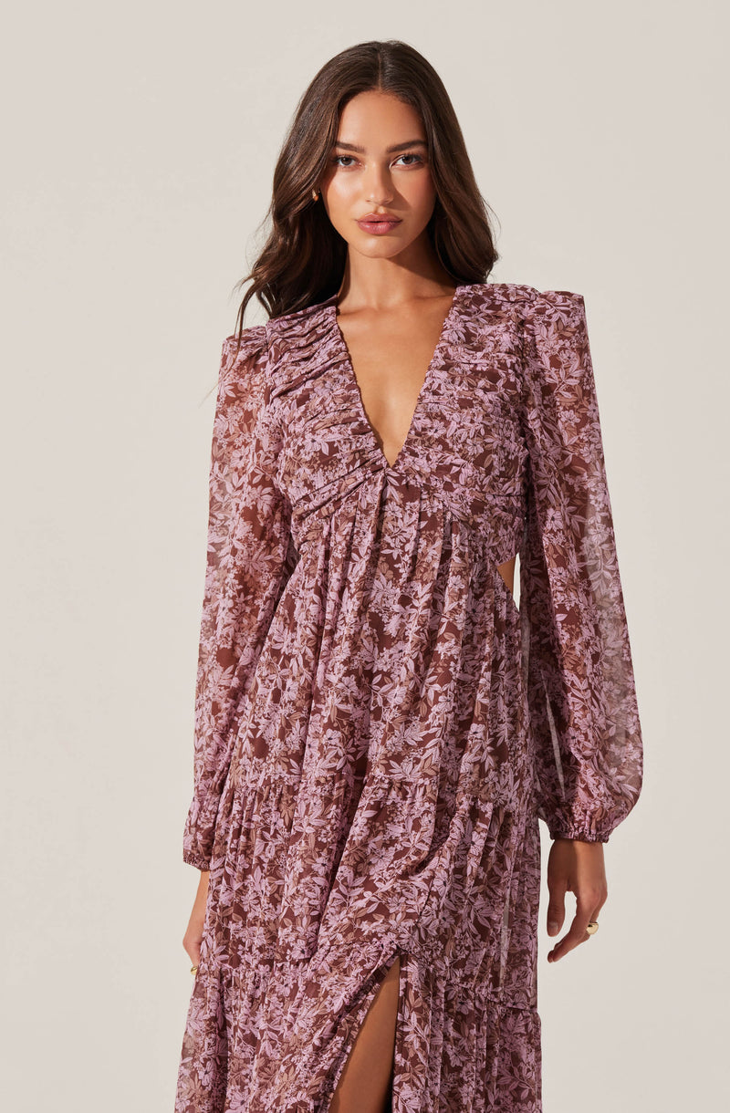 A woman is wearing a long, flowy dress with a floral print in shades of pink and mauve. The dress features a deep V-neckline, puffed long sleeves, and a slit up the front. The fabric appears lightweight and slightly textured, creating an elegant and romantic look.