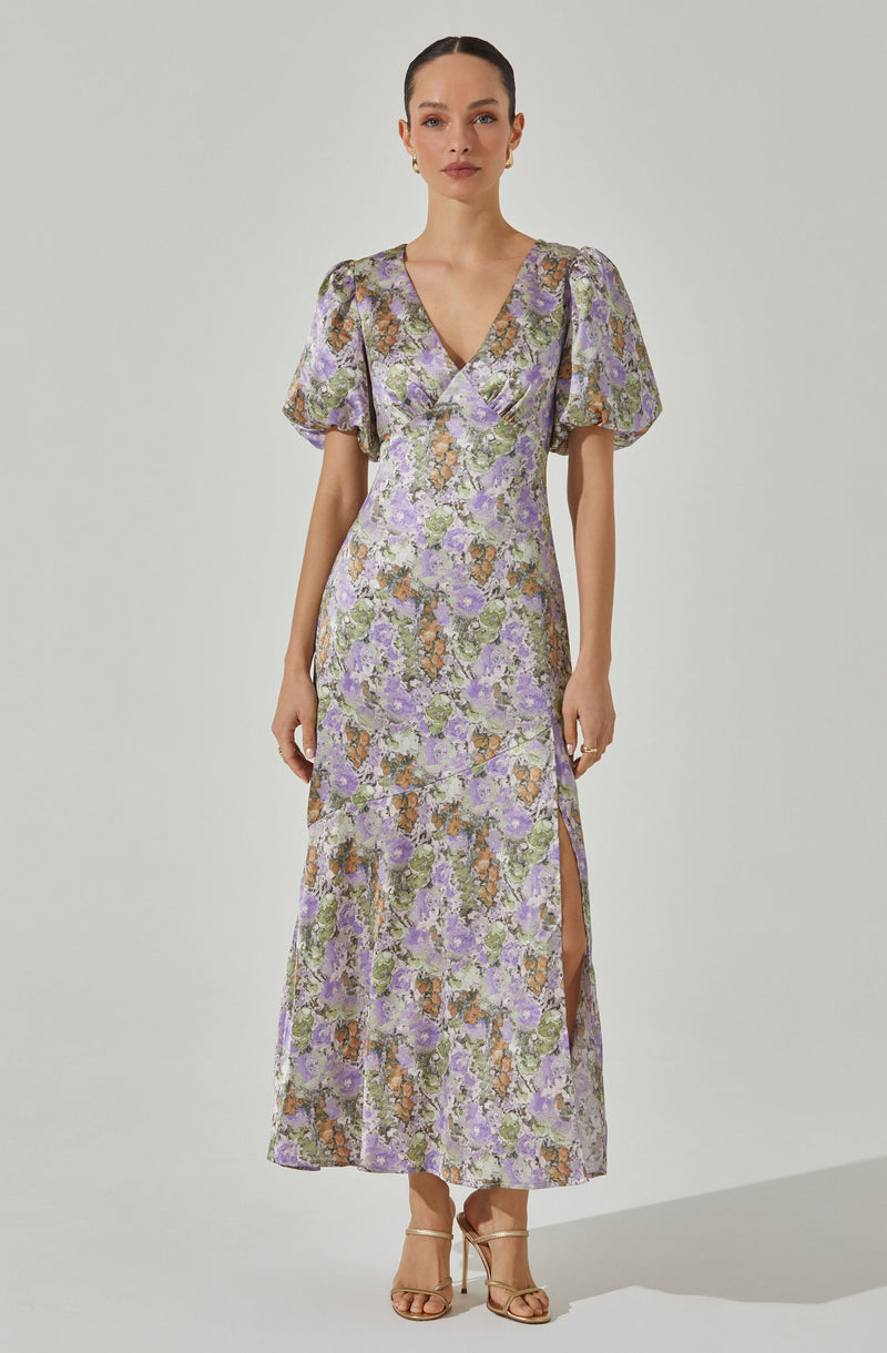 A woman is wearing a long, floral-patterned dress with a V-neckline. The dress features short, puffed sleeves and a side slit that extends up to her thigh. The fabric is decorated with a multicolored floral print in shades of purple, green, orange, and beige.
