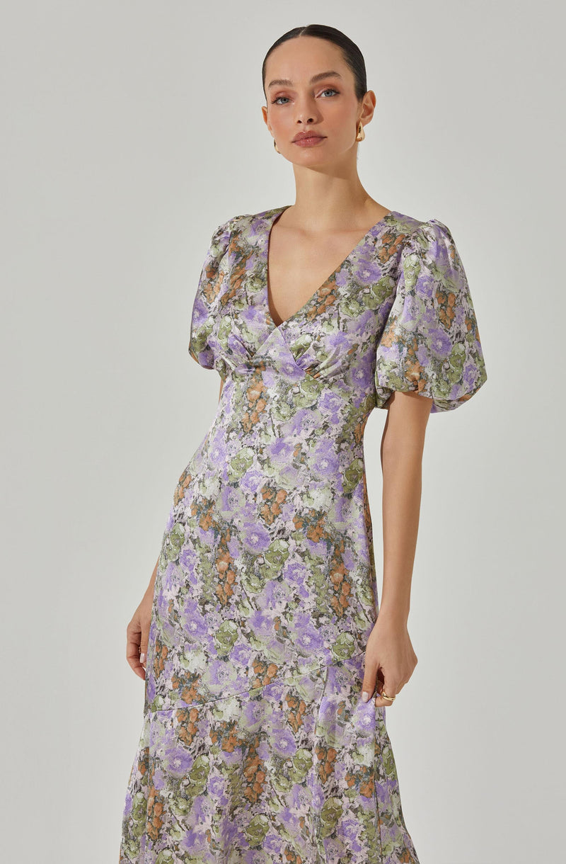 A woman is wearing a long, floral dress with a fitted bodice and a V-neckline. The dress features short, puffed sleeves and a pattern of purple, green, orange, and white flowers. The fabric appears to have a subtle sheen, and the dress has a slightly gathered waist that flows into a full-length skirt.