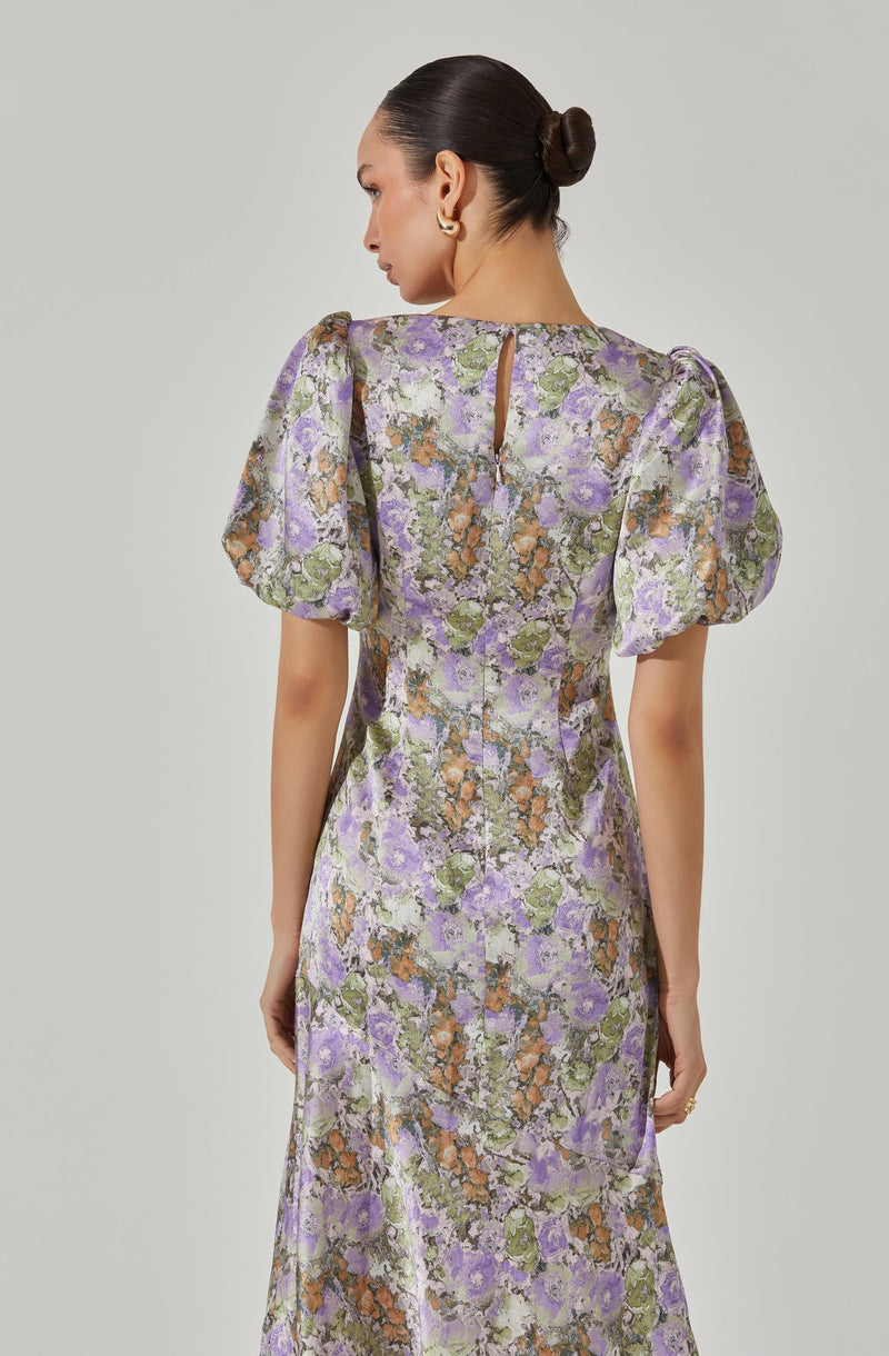 A woman is wearing a long dress with a floral pattern in shades of purple, green, orange, and cream. The dress features puffed short sleeves and a small keyhole opening at the back near the neckline.