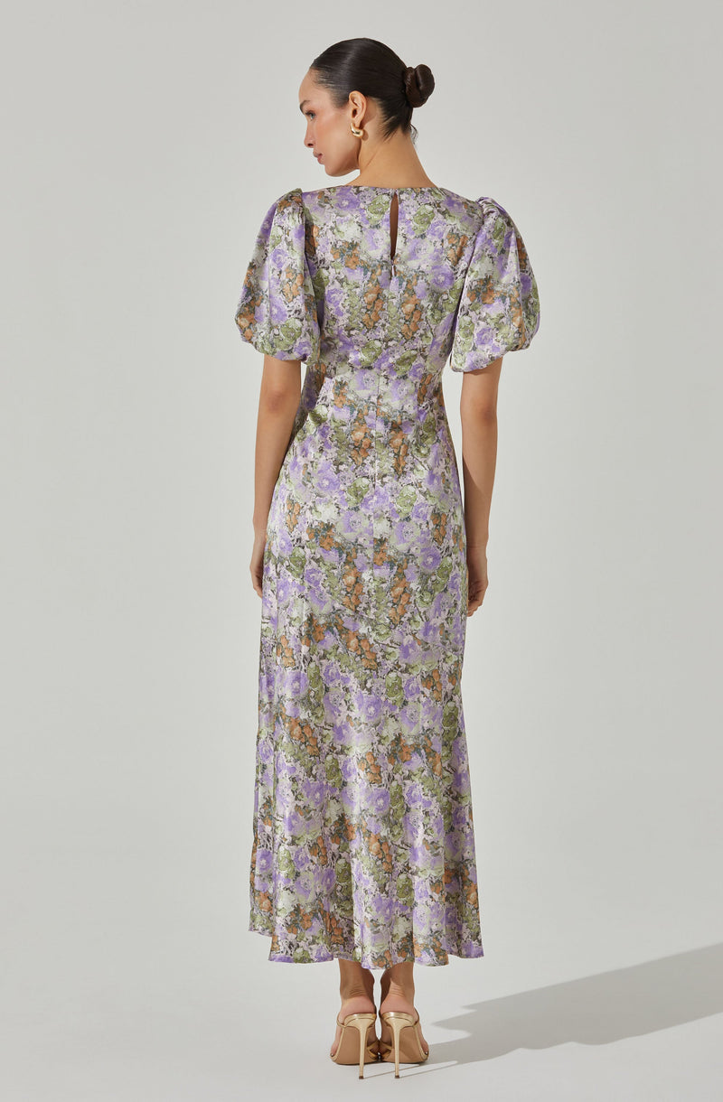 The dress is a full-length gown with a floral pattern featuring shades of purple, lavender, green, and brown. It has a fitted waist, a small keyhole opening at the back neckline, and puffed short sleeves. The fabric appears textured and slightly shiny, giving it an elegant appearance. The woman is wearing beige high-heeled shoes and has her hair styled in a sleek bun.