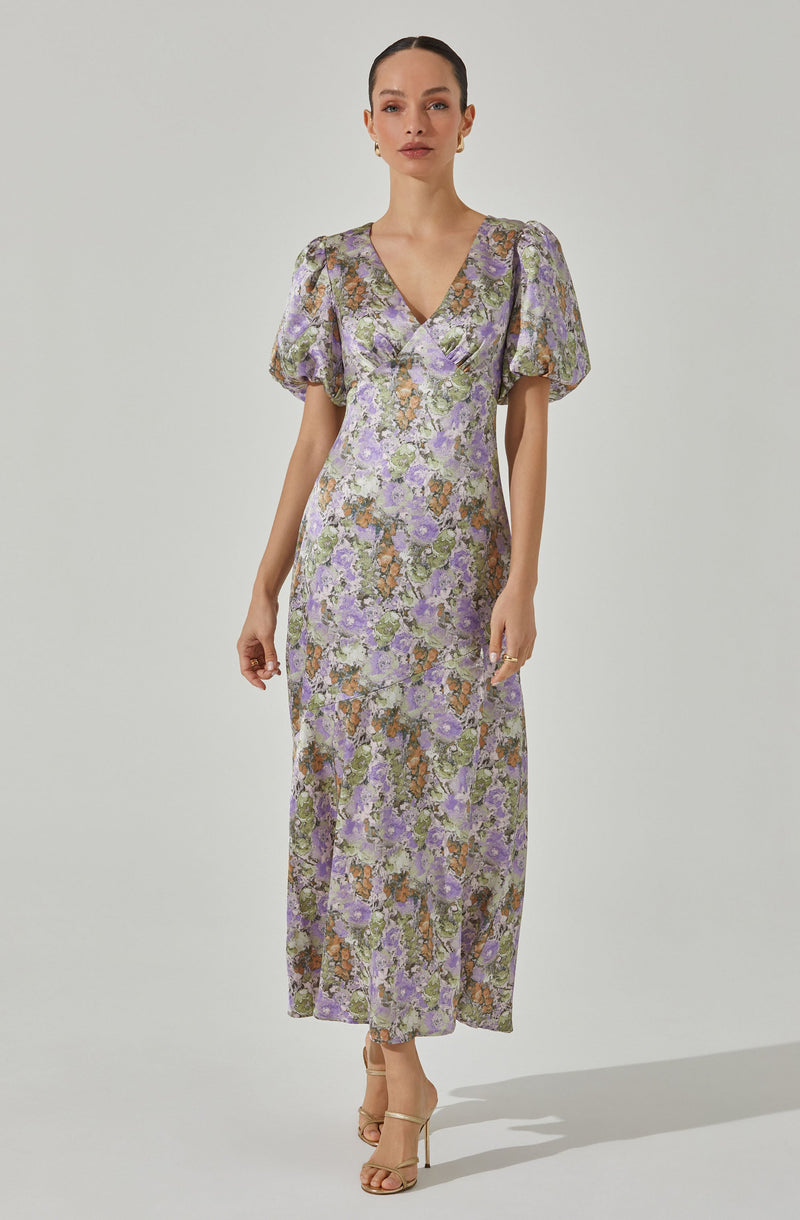 A woman is wearing a full-length dress with a floral print in shades of purple, green, and brown. The dress features short, puffed sleeves, a V-neckline, and a fitted waist. The fabric appears lightweight and slightly sheer.