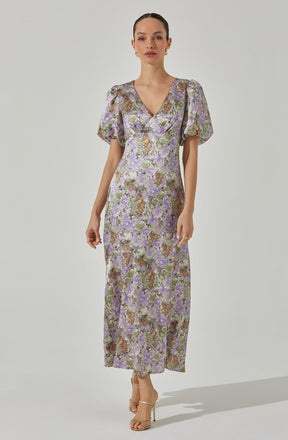 A woman is wearing a full-length dress with a floral print in shades of purple, green, and brown. The dress features short, puffed sleeves, a V-neckline, and a fitted waist. The fabric appears lightweight and slightly sheer.