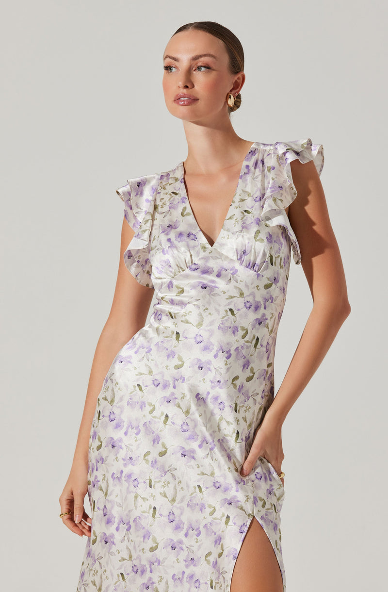 A woman is wearing a sleeveless, midi-length dress with a floral pattern in shades of purple and green on a light, possibly satin or silk fabric. The dress has ruffled cap sleeves, a V-neckline, and a side slit. She is accessorized with large, circular earrings and a ring, and has her hair pulled back.
