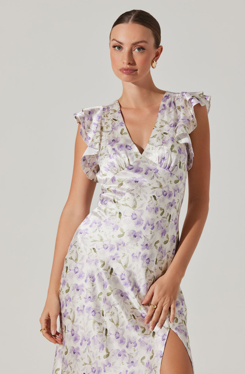A woman is wearing a sleeveless, satin-like dress with a floral pattern in shades of purple, lavender, and green on a white background. The dress features ruffled cap sleeves, a V-neckline, and a side slit.