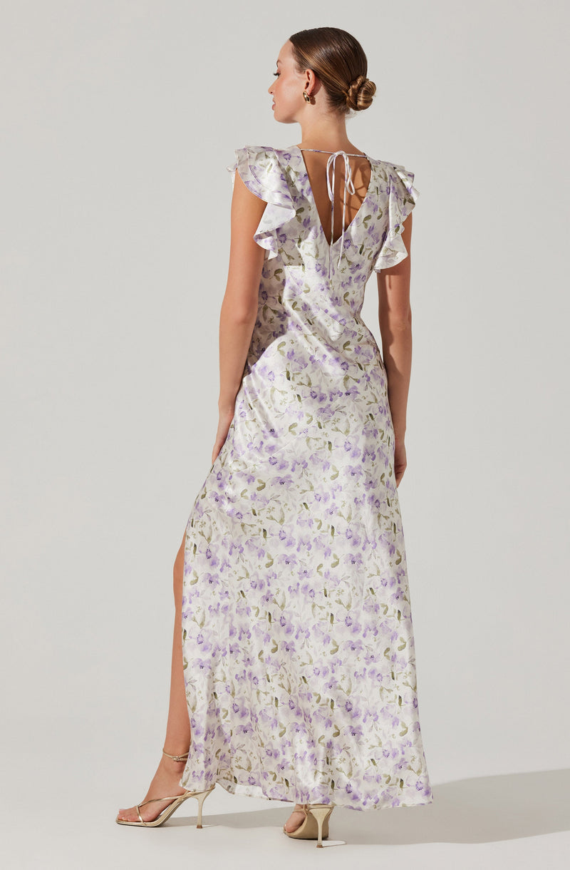 A woman is wearing a long, flowing dress with a light background and a floral pattern featuring shades of purple and green. The dress has ruffled cap sleeves, a V-shaped open back with a thin tie at the top, and a slit on the side. She is also wearing high-heeled sandals.