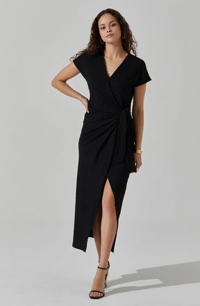 A woman is wearing a black dress with short sleeves and a V-neckline. The dress features a tied waist with a draped detail on one side, creating a slit that reveals her leg. She is also wearing black high-heeled sandals and gold jewelry, including earrings and stacked bracelets.