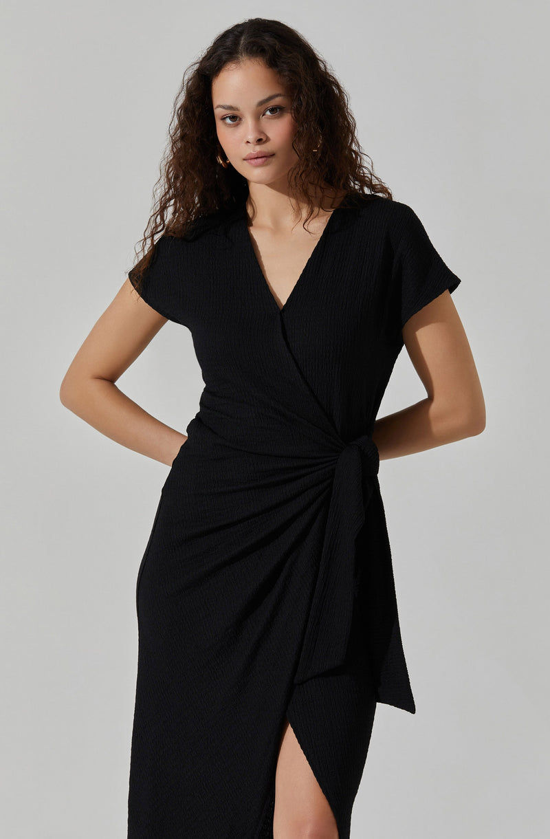 A woman wearing a black, short-sleeved dress with a V neckline. The dress features a knot at the waist, creating a gathered effect, and has a slit on the side that reveals part of her leg. The fabric appears textured with a subtle vertical rib pattern.