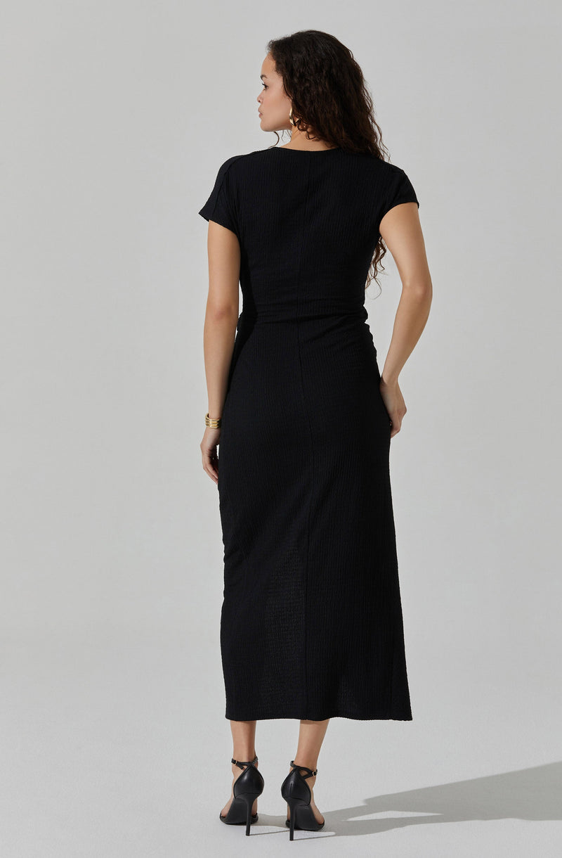A woman is wearing a fitted black short-sleeve top and a long, black, ribbed skirt that reaches her ankles. She has paired the outfit with high-heeled black shoes and gold jewelry, including bracelets. The overall look is elegant and monochromatic.