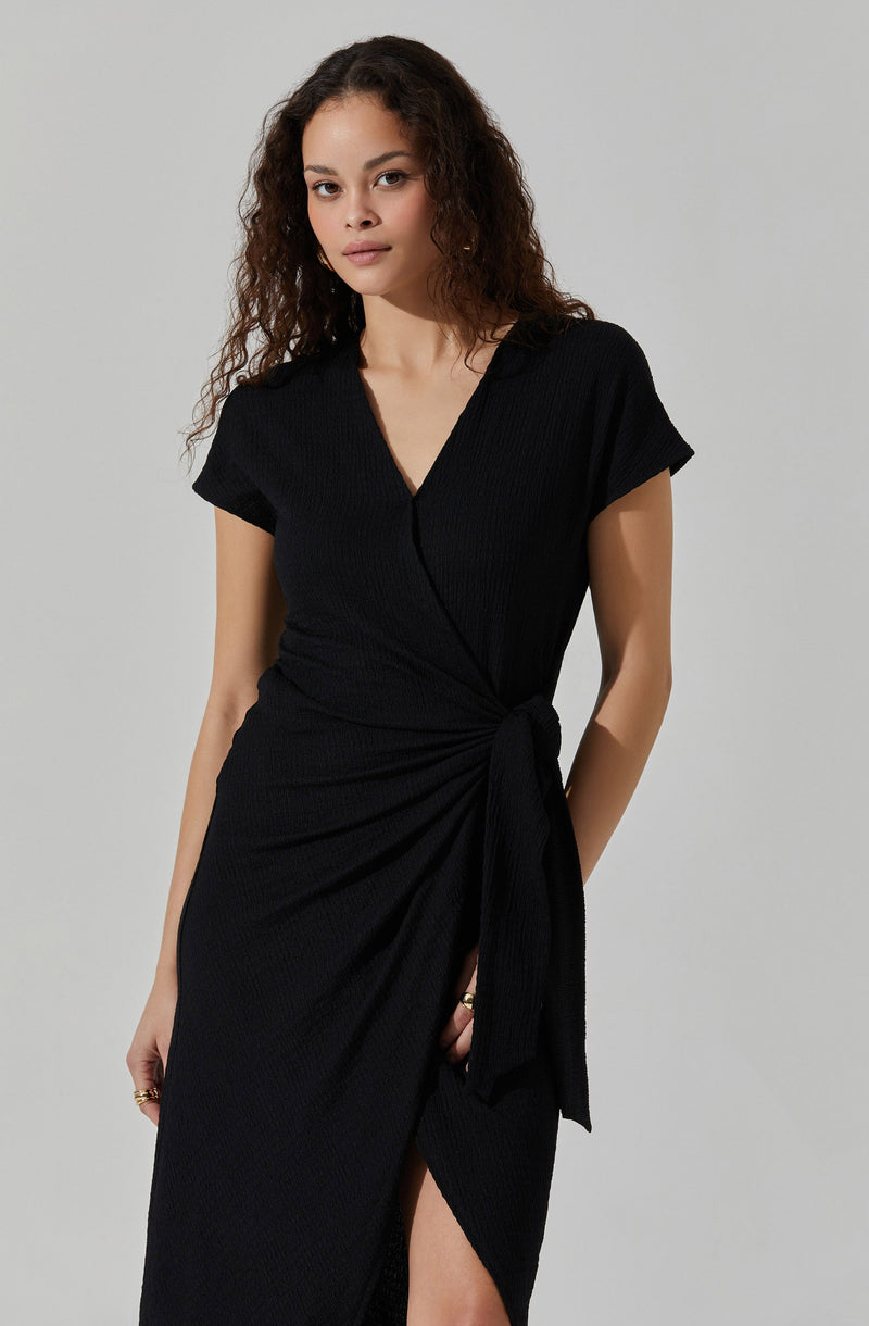 A woman is wearing a black wrap dress with a V-neckline and short sleeves. The dress features a tied detail at the waist and a slit up the front. The fabric has a textured pattern.