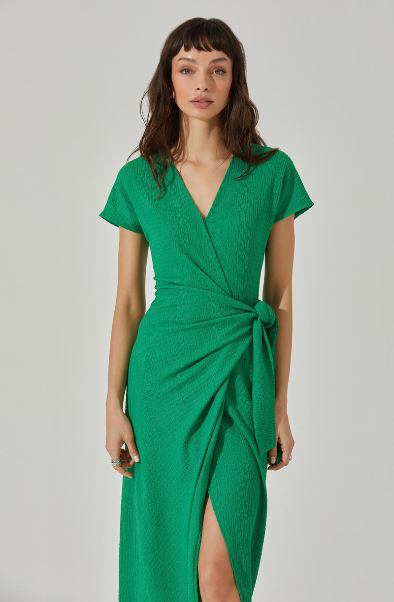 A woman is wearing a bright green dress with a textured fabric. The dress features a V-neckline, short sleeves, a fitted waist, and a side slit. It has a wrap-style design with a knot at the waist, creating a draped effect.