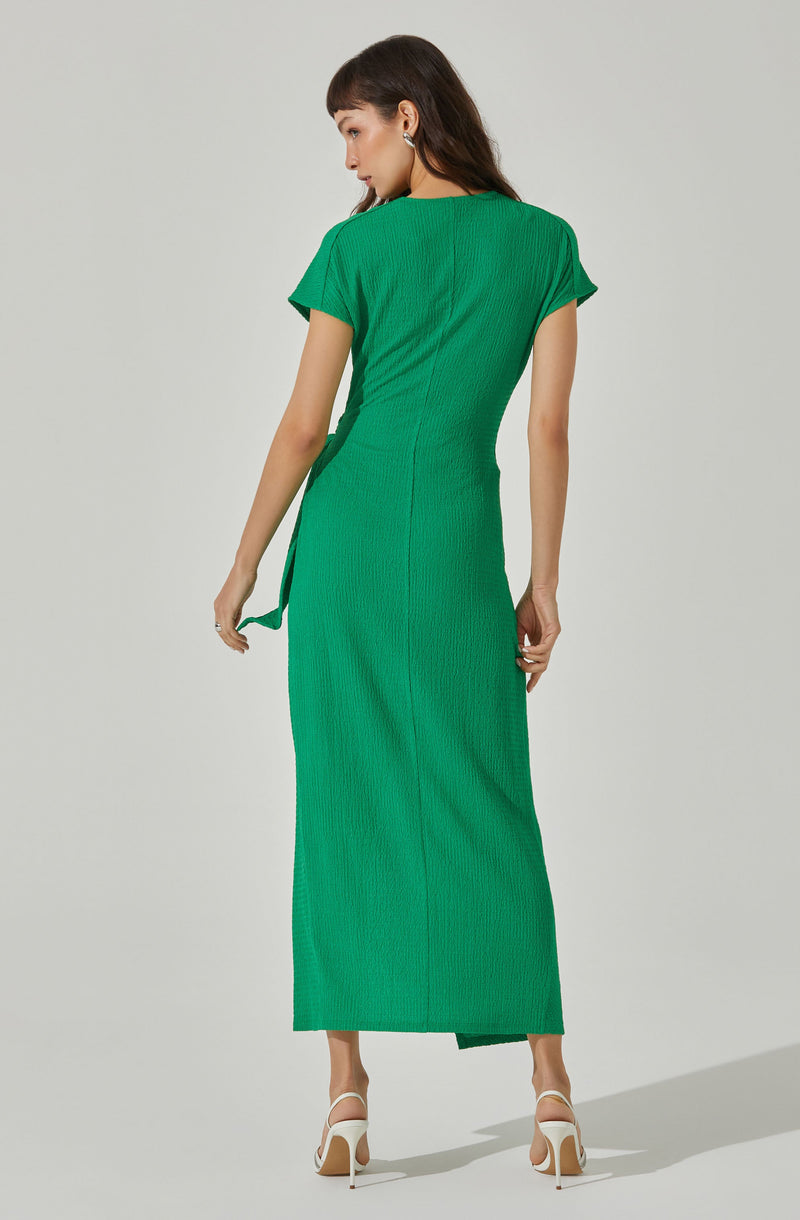 A woman is wearing a bright green textured dress with a fitted silhouette, short sleeves, and side slits at the hem. The dress has a smooth, slightly shiny finish. She pairs the dress with white high-heeled sandals and silver earrings.