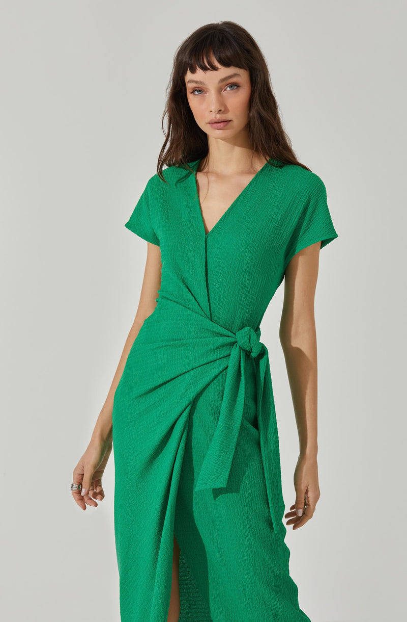 A woman is wearing a bright green dress made of textured fabric. The dress features a V-neckline and short sleeves. It has a waist tie that is knotted on the right side, creating a draped effect with pleats falling from the knot.