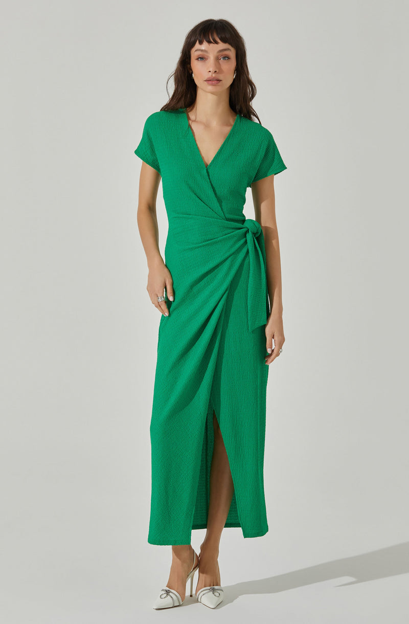 A woman is wearing a vibrant green dress with a wrap-style bodice, short sleeves, and a matching fabric belt tied at the waist. The dress has a textured fabric and a slit at the front, revealing her legs. She is also wearing beige high-heeled shoes with a bow detail on the front.