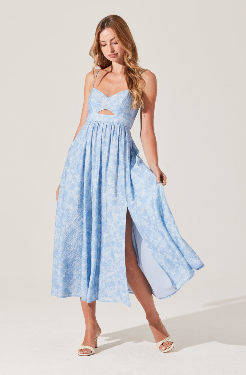 A woman is wearing a light blue, sleeveless maxi dress with spaghetti straps. The dress features a subtle floral pattern, a cut-out detail at the chest, and a high slit on one side revealing her leg. She is also wearing beige high-heeled shoes.