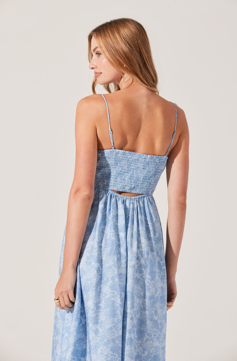 A woman with light brown hair is wearing a light blue, sleeveless dress with thin spaghetti straps. The dress features a fitted, textured bodice with a small cutout at the back, and a gathered, flowing skirt made of a slightly shiny, patterned fabric.