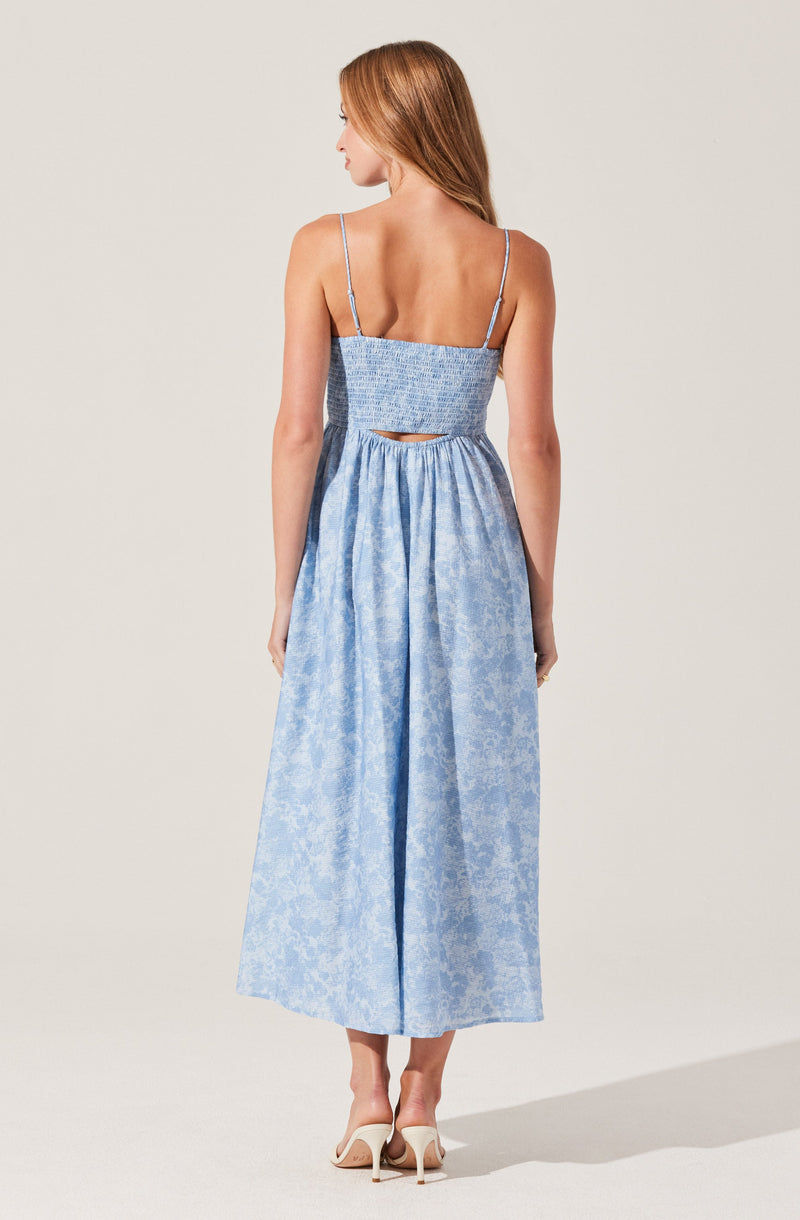 A woman is wearing a light blue, floral-patterned dress with thin spaghetti straps. The dress has a smocked bodice with a small cut-out detail at the back, and a high-waisted, full-length skirt with a subtle floral texture. She is also wearing white high-heeled shoes.