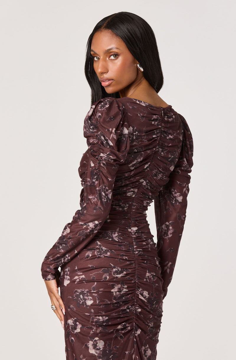 A woman is wearing a long-sleeved, fitted dress with a dark maroon color and an abstract, floral print design. The dress features ruched detailing along the back and sides, creating a textured, gathered appearance. The fabric appears shiny and smooth.
