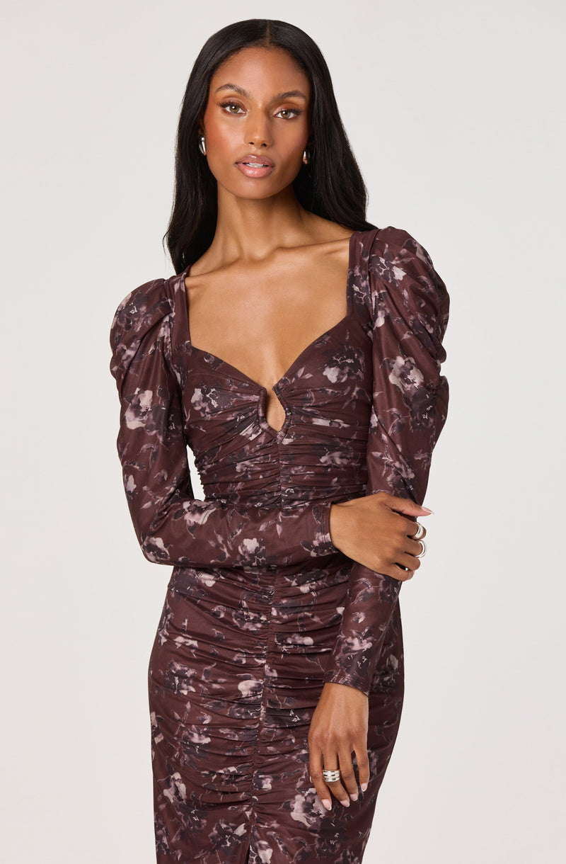 A woman is wearing a long-sleeved, fitted dress with a brown and pink floral pattern. The dress features ruched detailing along the front and has a keyhole cutout at the chest with a small twist knot in the center. It also has puffed shoulders and a scoop neckline, creating a stylish and elegant look.