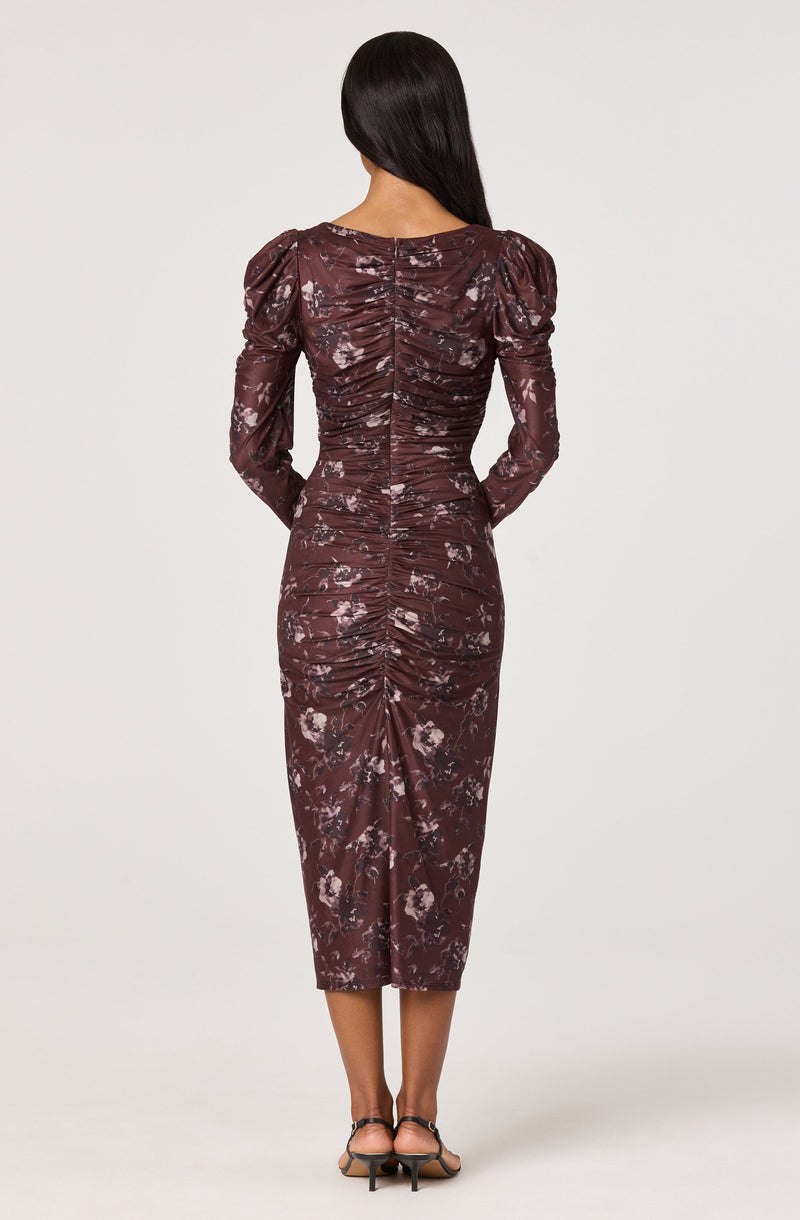 A woman is wearing a fitted, long-sleeved dress with a dark maroon base color and a floral pattern in shades of pink and white. The dress features ruched detailing along the center back and down the length of the skirt, with puffed shoulders adding volume to the sleeves. She is also wearing black open-toe heels.