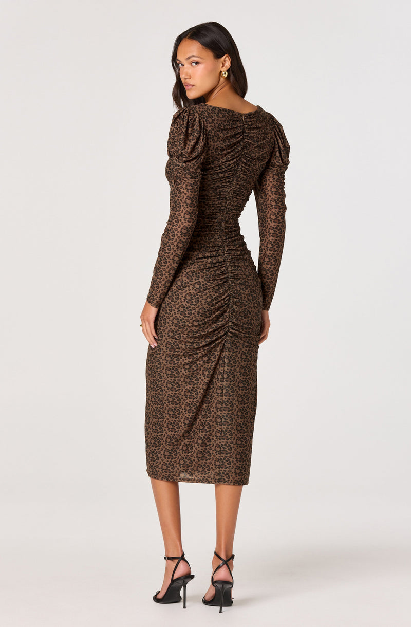 A woman is wearing a long, fitted dress with a leopard print pattern. The dress features long, sheer sleeves and a ruched design along the back and center front, creating a textured, gathered look. The dress extends to mid-calf length and has a sleek, elegant silhouette. She completes the outfit with black high-heeled sandals.