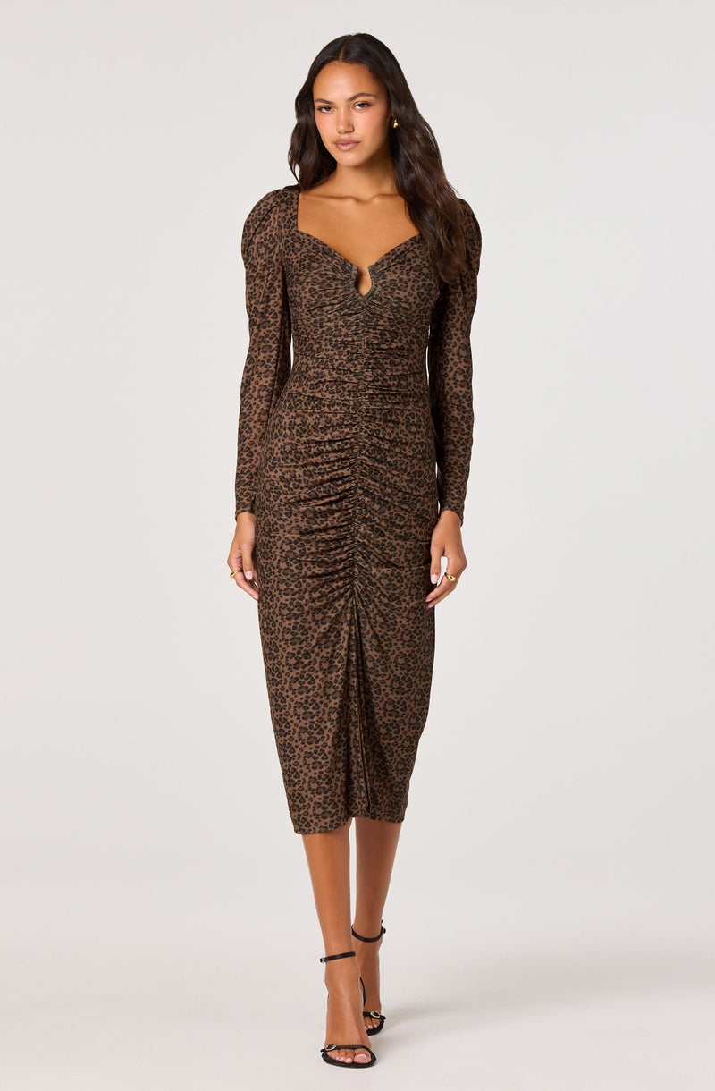 A woman is wearing a long, fitted dress with a leopard print pattern in shades of brown and black. The dress has long, sheer sleeves and a sweetheart neckline with a small keyhole detail. The dress features ruched gathering down the front center, creating a draped effect. The hem of the dress falls below the knee. She is also wearing black high-heeled sandals with ankle straps.