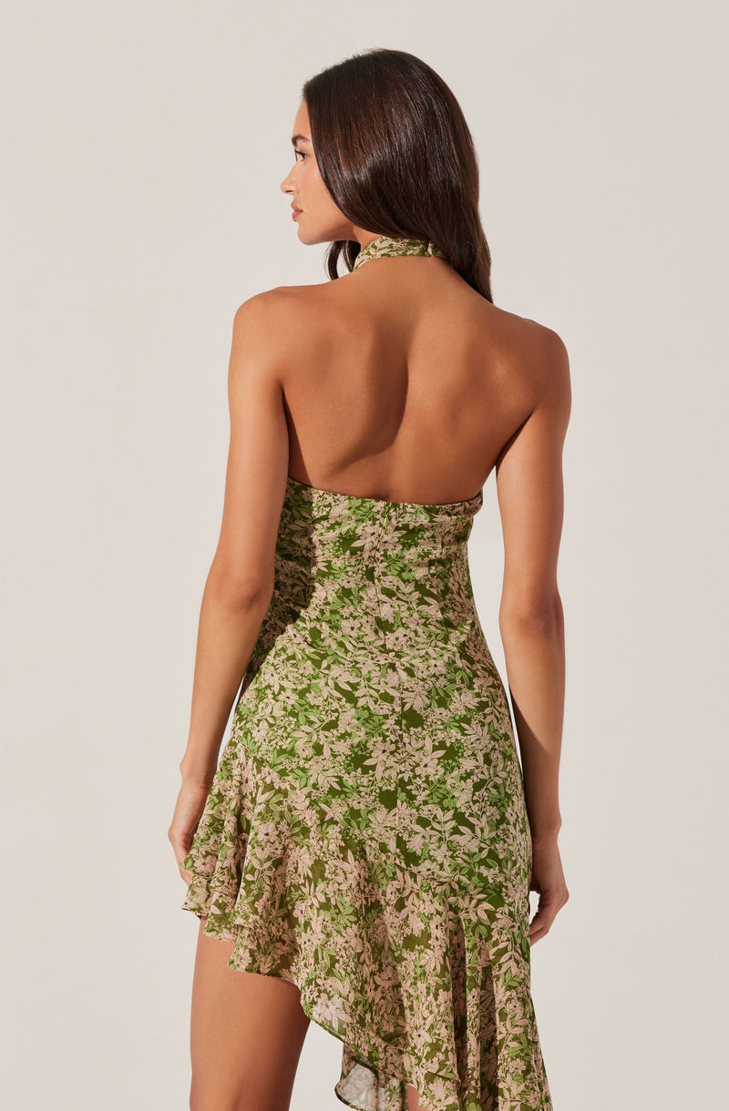 A woman is wearing a sleeveless dress with a halter neckline. The dress features a floral pattern in shades of green and light pink, with an asymmetrical ruffled hem. The back of the dress is open, revealing her bare shoulders and upper back.